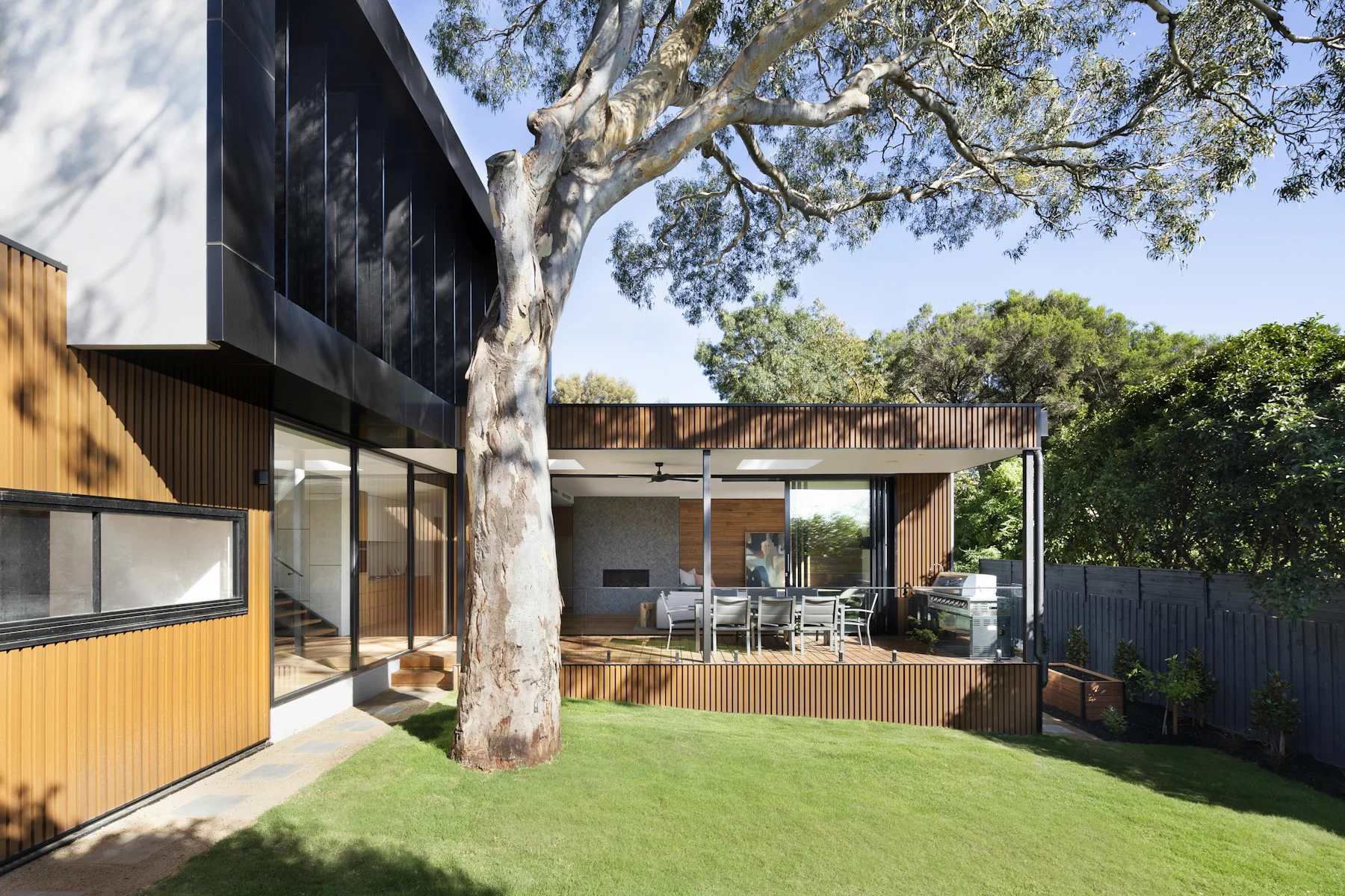 Exterior de una vivienda contemporánea con jardín, gran árbol y terraza cubierta con comedor